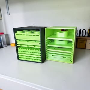 May include: Two storage organizers, one black and one lime green, sit side-by-side. Each has multiple shelves and compartments. The lime green organizer has a matching green interior. The black organizer has a green interior. Both are on a white surface.