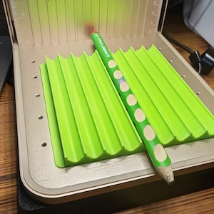 May include: A green pencil with white polka dots rests on a bright green, corrugated surface inside a laser engraving machine. The machine has a metallic gold finish and a fan at the top.