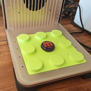 May include: A silver and black laser engraving machine with a green mat and a bottle cap. The bottle cap has a purple and orange design. The machine has a fan and measurement markings.