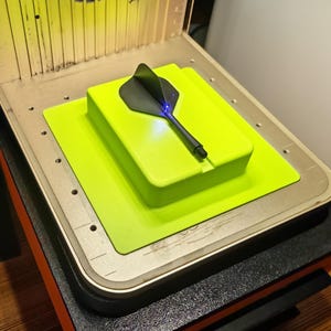 Könnte beinhalten: Ein 3D-Drucker mit einem schwarzen und grauen pfeilförmigen Objekt auf einer neongrünen Plattform. Der Drucker hat eine orangefarbene Basis und eine transparente Oberseite mit einem Licht im Inneren. Das Objekt ist zentriert.
