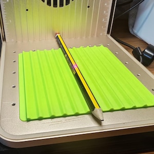 May include: A pencil with black and yellow stripes rests on a bright green, ridged mat inside a silver machine. The machine has a fan and a ruler-like scale on the side. The pencil's tip is pointed towards the machine's interior.