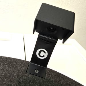 Könnte beinhalten: Eine schwarze Kamera mit quadratischem Gehäuse und einem Scharnierhalter. Der Halter hat ein weißes "C"-Logo. Die Kamera ist auf einer dunkelgrauen, strukturierten Oberfläche montiert, wahrscheinlich ein Helm oder ein ähnlicher Artikel.