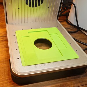 May include: A silver-colored laser engraving machine with a bright green insert and a circular cutout. The machine has a fan and a ruler marked in inches. The device sits on a wooden surface.