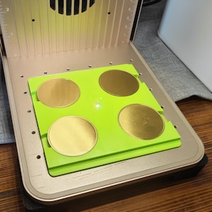 May include: A gold and green laser engraving machine with a fan and a green platform holding four gold-colored circular objects. The machine has a metallic finish and is set on a wooden surface.