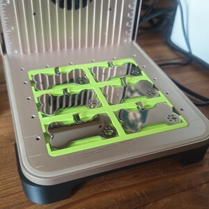 May include: A silver-colored metal device with a green insert holding several dog bone-shaped metal tags. The device has a fan and vertical lines on the top. The tags have a small loop for attaching to a collar.