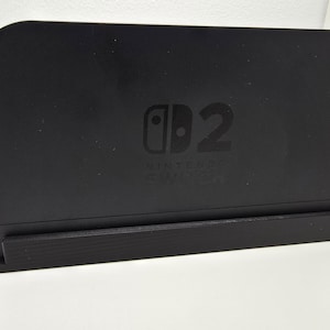 Könnte beinhalten: Schwarze Nintendo Switch-Dockingstation mit dem Nintendo Switch-Logo und der Zahl 2 auf der Vorderseite. Die Dockingstation ist rechteckig mit leicht gebogener Oberseite und einem Sockel.