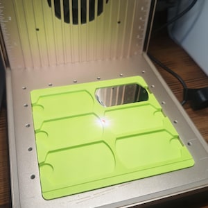 May include: A 3D printer with a bright green platform and a reflective surface. The printer has a yellow-tinted cover and a fan. The platform has a series of rectangular cutouts.