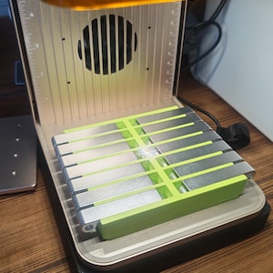 May include: A 3D printer with a silver and green build plate. The printer has a clear orange top and a silver body with a fan and measurement markings. The build plate is composed of silver metal pieces set in a green frame.