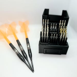 Könnte beinhalten: Drei schwarze Darts mit orangefarbenen Flights, ein schwarzes Dart-Etui mit einer Vielzahl von Metallschafts.