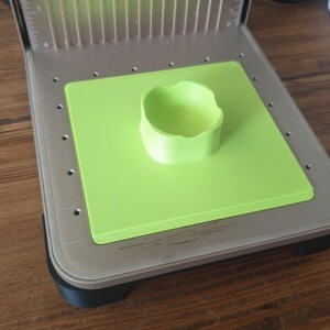 Könnte beinhalten: Eine Lasergravurmaschine mit einer limettengrünen Basis und einem passenden zylindrischen Objekt. Die Maschine hat einen silbernen Metallrahmen mit einem Lüfter und Messmarkierungen. Das Objekt ist auf der grünen Basis zentriert.
