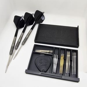 Könnte beinhalten: Ein schwarzes Dart-Set mit drei Darts, einer Aufbewahrungsschale und zusätzlichen Dart-Komponenten. Die Darts haben schwarze Flights, silberne Schäfte und silberne Spitzen. Das Set enthält eine schwarze Schale mit dem "TARGET"-Logo.