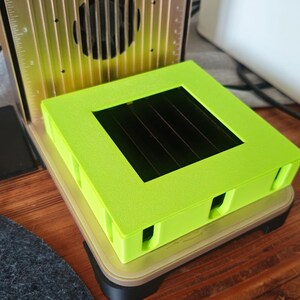 May include: A lime green square tray with a rectangular opening sits atop a beige and black base. The tray is part of a larger machine with a metallic gold and black finish. The machine has a series of vertical lines and a fan.