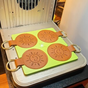 May include: Four brown leather keychains with a sun design and gold-colored rings are arranged on a light green surface. The keychains are 50 mm in diameter. The keychains are in a laser engraving machine.