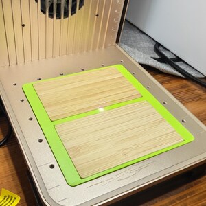 May include: A laser engraving machine with a gold-colored metal casing. Inside, two rectangular bamboo pieces sit on a green mat. A laser beam is visible, and a fan is present. The machine has a measurement scale.