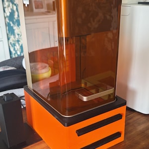 Può includere: Stampante 3D arancione e marrone con camera di resina trasparente color ambra. La stampante ha una base rettangolare con due barre orizzontali nere e un'etichetta di avvertimento. È visibile un pulsante di arresto rosso.