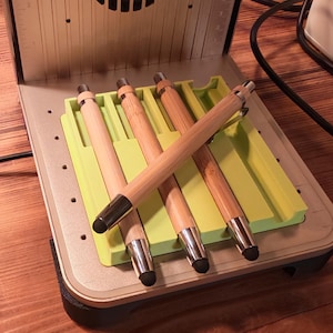 May include: A collection of bamboo pens with silver accents and black stylus tips, resting on a lime green tray. The pens are displayed inside a metallic device with a vent design.