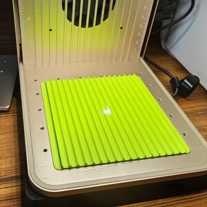 May include: A 3D printer with a bright green, ridged platform inside a metallic enclosure. The printer has a fan and a clear orange top. The platform is illuminated by a bright light.