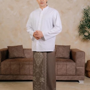 May include: A white long-sleeved shirt with a mandarin collar and subtle embroidery on the chest, paired with a brown sarong featuring a geometric pattern and the text ADYATMA printed near the bottom hem, worn with black open-toe sandals.