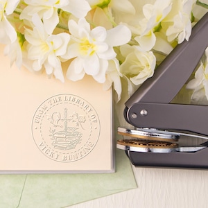 May include: A silver self-inking stamp with a round design featuring a stack of books, a sword, and a dragon. The text around the design reads "From the Library of Vicky Buntine".