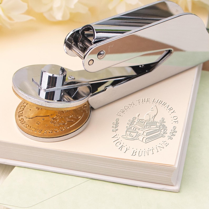 May include: A silver self-inking stamp with a round, gold-colored stamp pad. The stamp is imprinted with the text "From the Library of Vicky Buntine" and a design of a cat sitting on a stack of books.