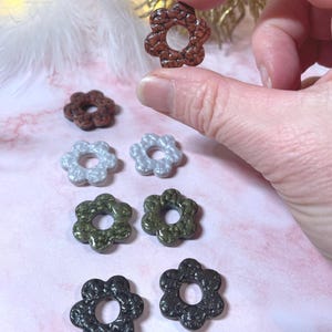 May include: A collection of flower-shaped beads in various colors, including brown, silver, olive green, and black. The beads have a textured surface and a hole in the center. One bead is held between fingers.