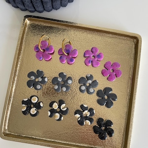 Peut inclure: Une collection de boucles d'oreilles en forme de fleurs de différentes couleurs, dont le violet, le noir et l'or, présentées sur un plateau doré. Certaines boucles d'oreilles ont des cerceaux dorés. Les boucles d'oreilles sont disposées en grille.