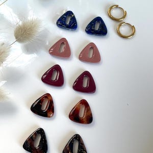 Puede incluir: Una colección de pendientes en varios colores y estilos. Hay seis pares de pendientes de botón triangulares en azul marino, rosa, burdeos y patrones de carey. También se incluyen dos pequeños pendientes de aro dorados. Los pendientes se muestran sobre una superficie blanca.