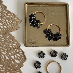 Peut inclure: Boucles d'oreilles créoles dorées avec des détails floraux noirs, présentées sur un plateau doré. Des perles noires en forme de fleurs et un seul anneau doré sont également visibles.