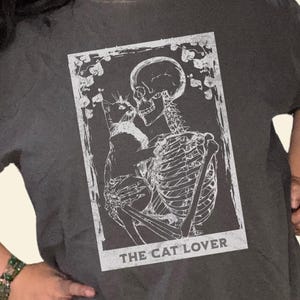 Puede incluir: Camiseta gris oscuro con un gr&aacute;fico blanco de un esqueleto abrazando a un gato, con el texto "THE CAT LOVER". La camiseta tambi&eacute;n tiene el texto "Fur Baby Lovers" y un logotipo. La camiseta tiene un corte holgado y de gran tama&ntilde;o.