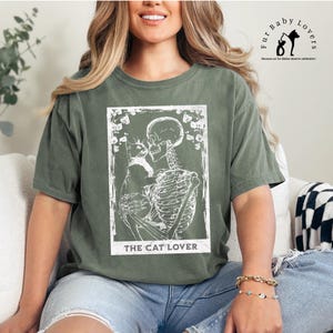 Puede incluir: Camiseta verde oliva con un gr&aacute;fico blanco de un esqueleto abrazando a un gato, con el texto "THE CAT LOVER" debajo. La camiseta tiene un corte holgado y est&aacute; hecha de un material suave. Parte de la colecci&oacute;n Fur Baby Lovers.