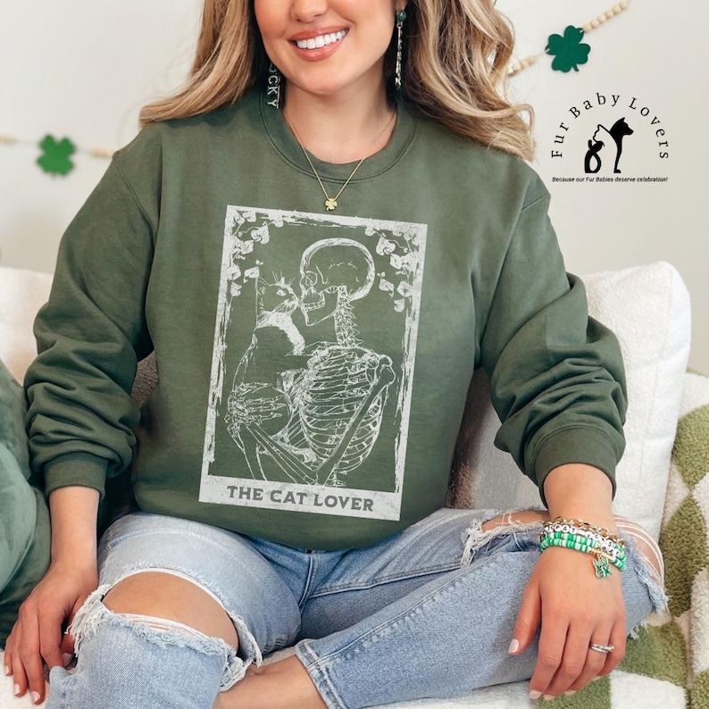 Puede incluir: Una sudadera verde oliva con un gr&aacute;fico blanco de un esqueleto sosteniendo un gato, con el texto "THE CAT LOVER" debajo. La sudadera tiene cuello redondo y mangas largas. La imagen tambi&eacute;n incluye el logo "Fur Baby Lovers".