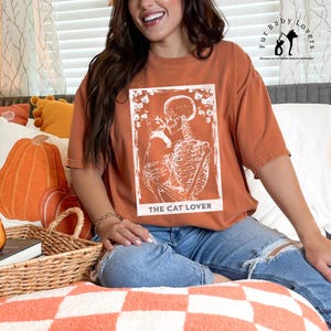Puede incluir: Camiseta de color naranja quemado con un gr&aacute;fico blanco de un esqueleto abrazando a un gato, con el texto "THE CAT LOVER" en la parte inferior. La camiseta tiene mangas cortas y un corte holgado. La imagen tambi&eacute;n incluye el texto "Fur Baby Lovers" y un logotipo.