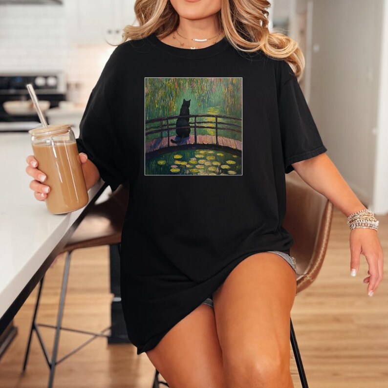 May include: A black t-shirt with a graphic print of a black cat sitting on a bridge over a pond with water lilies. The pond is surrounded by trees and the sky is a light blue.