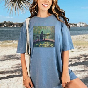 May include: A light blue oversized t-shirt with a graphic print of a black cat sitting on a bridge over a pond with water lilies. The pond is surrounded by green trees and the sky is blue.