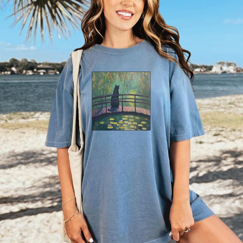 May include: A light blue oversized t-shirt with a graphic print of a black cat sitting on a bridge over a pond with water lilies. The pond is surrounded by green trees and the sky is blue.