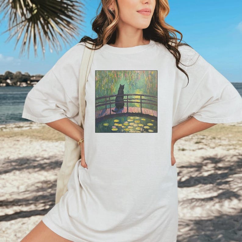 May include: A white t-shirt with a graphic print of a black cat sitting on a bridge over a pond with water lilies. The pond is surrounded by green trees and the sky is blue.