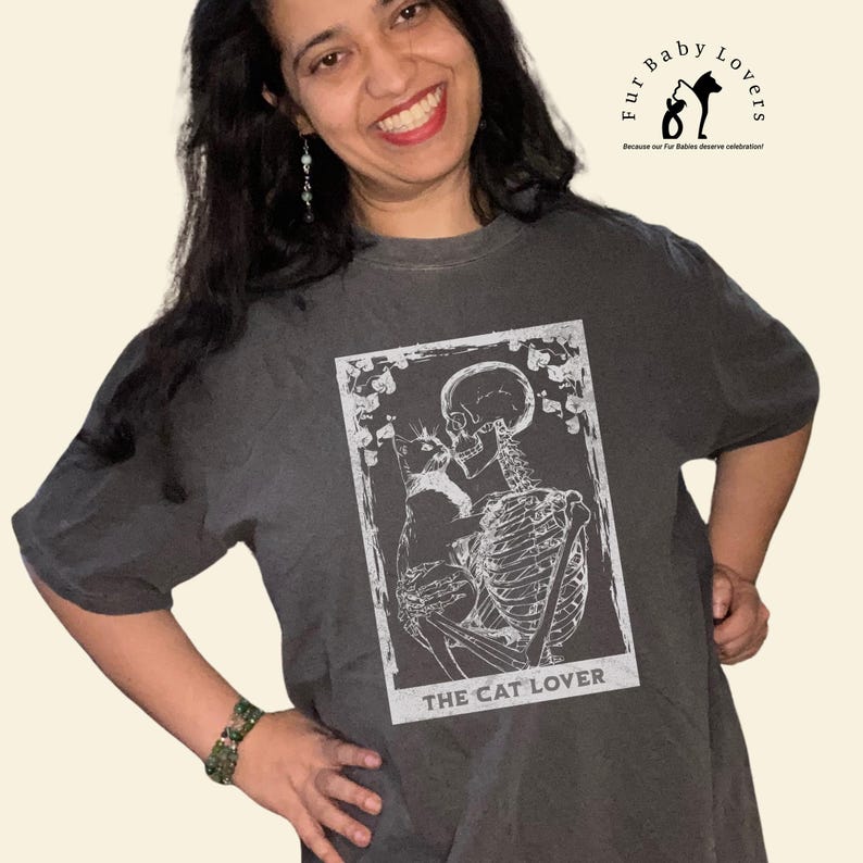 Puede incluir: Camiseta gris oscuro con un gr&aacute;fico blanco de un esqueleto abrazando a un gato, con el texto "THE CAT LOVER". La camiseta tambi&eacute;n tiene el texto "Fur Baby Lovers" y un logotipo. La camiseta tiene un corte holgado y de gran tama&ntilde;o.
