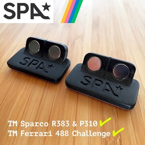 Puede incluir: Dos soportes negros impresos en 3D con el texto SPA en relieve y un símbolo de estrella. Cada soporte tiene dos imanes metálicos circulares integrados en la cara vertical. El texto TM Sparco R383 & P310 y TM Ferrari 488 Challenge es visible.