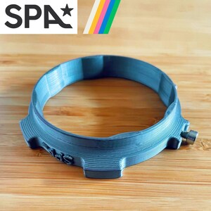 May include: A gray, circular object with a screw, possibly a watch component or a similar accessory. The word "SPA" is visible in the upper left corner, along with a colorful striped design. The object is on a wooden surface.
