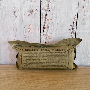 May include: A rectangular olive-drab fabric packet labeled Dressing, Shell, Mark III. The text provides instructions for use, including how to tear the cover, extract the dressing, and apply the bandage to a wound. The packet is sealed.