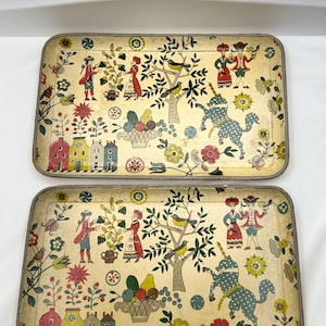 May include: Two rectangular serving trays with a vintage design. The trays feature a cream-colored background with a colorful folk art scene, including figures, trees, birds, and a whimsical blue animal. The trays have a metallic silver trim.