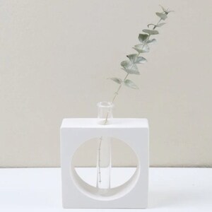 May include: White ceramic vase with a circular cutout and a clear glass test tube inside. A sprig of green eucalyptus is in the test tube.