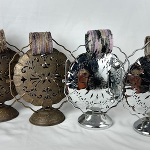 May include: Four vintage metal display stands with ornate cutouts. Each stand features a round, openwork top with a decorative base. The stands are holding colorful beaded bracelets.