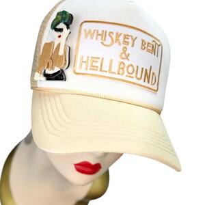 May include: Cream-coloured trucker hat with a white front panel featuring the text "WHISKEY BENT & HELLBOUND" in gold lettering. An acrylic pin of a woman in sunglasses is on the side. The hat has a curved brim and mesh back.