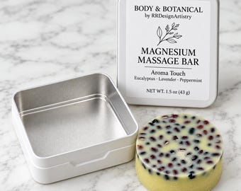 Puede incluir: Una barra de masaje de magnesio redonda con inclusiones oscuras similares a bayas junto a una lata de metal plateado abierta. La tapa tiene una etiqueta blanca que dice BODY & BOTANICAL, MAGNESIUM MASSAGE BAR, 43 g.