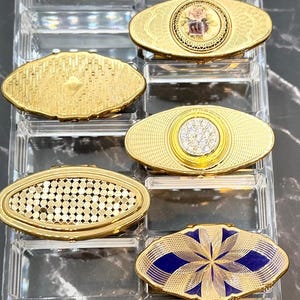 May include: A collection of vintage gold compact mirrors in various oval designs. Some feature intricate patterns, floral accents, or jewel-like embellishments. The compacts are displayed in a clear, segmented organizer.