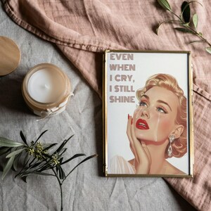 May include: A framed print featuring a vintage-style illustration of a woman with red lipstick and tears running down her cheeks. The text on the print reads "Even when I cry, I still shine."