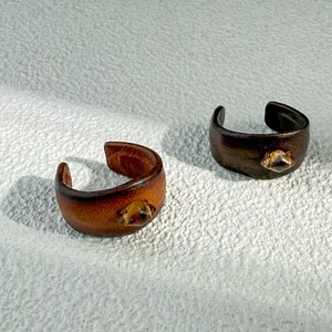 Puede incluir: Dos anillos de cuero hechos a mano. Uno es de color marrón claro y el otro de color marrón oscuro. Cada anillo tiene una pequeña gema transparente. El diseño abierto permite un ajuste regulable. Los anillos son de cuero.