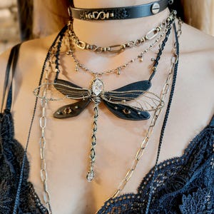 May include: A layered necklace set featuring a black leather choker, multiple chains, and a dragonfly pendant. The dragonfly pendant has black wings, a silver wire frame, and a central crystal. The chains are silver and gold-toned.