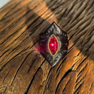 May include: A dark, shield-shaped ring with a large, oval, red gemstone at its center. The ring has intricate, embossed detailing and a gold-colored metal border around the gem. The ring is set against a textured, wooden surface.
